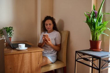 Bir kadın ev ortamında rahat bir şekilde oturur, bilgisayarla uğraşırken akıllı telefonunu kullanır. Bitkiler ve bir fincan kahve verimli bir uzaktan çalışma ortamı sunan rahat bir atmosfer ekler..