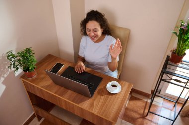 Sanal bir toplantı sırasında neşeli bir kadın bir fincan kahveyle rahat ofis masasına oturmuş el sallıyor..