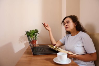 Bir kadın masa başında oturmuş uzaktan çalışıyor ve bir kitaptan bahsederken bilgisayarını kurcalıyor. Kurulumda bir kahve fincanı, yapraklı bir bitki ve rahat bir ev ofisinde iyi ışıklandırılmış bir çevre yer alıyor..