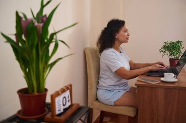 Bir kadın elinde dizüstü bilgisayarla masada oturuyor ve uzaktan çalışıyor. Etrafında saksı bitkileri ve düzenli bir alan var. Sakin ve üretken bir çalışma ortamını vurguluyorlar..