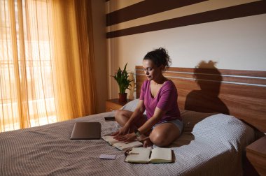Günlük kıyafetli bir kadın yatak odasında yazılı bir şekilde oturuyor. Sıcak güneş ışığı perdelerin arasından süzülerek dingin ve odaklanmış bir ortam yaratır..
