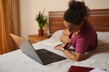 Dizüstü bilgisayarı ve not defteriyle yatakta yatan bir kadın akıllı saatini kontrol ediyor. Yatağın başucundaki masaya sıcak bir ortam sağlayan bir bitki yerleştiriliyor. Üretkenlik ve rahatlama sahnesi.