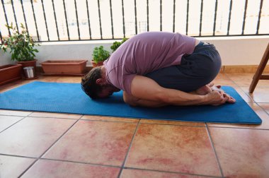 Bir adam, balkonundaki parlak mavi bir paspasın üzerinde rahatlatıcı bir yoga pozu veriyor. Etrafı saksı bitkileriyle çevrili, sakin bir ortamda rahatlık ve refahı kucaklıyor..