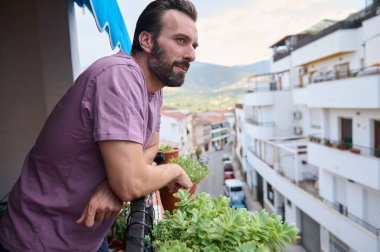 Evinin balkonuna yaslanmış, bitkilerle çevrili bir adam aşağıdaki manzaralı bir kasaba yolunu izliyor. Görüntü sakin bir şehir yaşam tarzını yakalıyor..