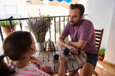 Bir baba ve genç bir kız, güneşli bir balkonda, kırsal bir ahşap masada, bir fincan kahve ve lavanta buketi eşliğinde, sıcaklığı ve aile bağlarını aktaran bir sohbete girişiyor..