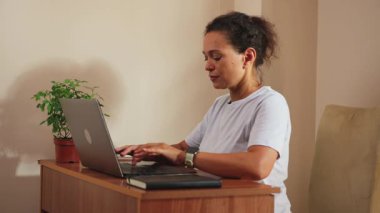 Odaklanmış bir kadın özenle ev ofisinden bilgisayarını açıp daktilo ederek çalışıyor. Verimli bir şekilde dijital teknolojiyi ve geleneksel aletleri kullanıyor, mesela bir defter, üretken bir uzaktan çalışma için. Bu sahne modern hayatın özünü yansıtıyor.