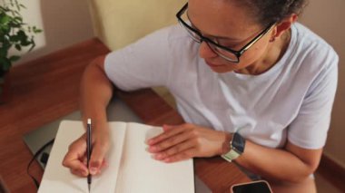 Odaklanmış bir kadın özenle not defterine kendi masasında yazıyor. Konsantrasyonu ve verimliliği somutlaştıran uzaktan çalışma ya da öğrenim görüyor. Bu sahne modern ev yaşamını ve kendini adamış öğrenmeyi ön plana çıkarıyor..