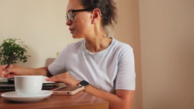 Gözlüklü odaklanmış bir kadın, özenle not defterine notlar yazarak ev ofisinde çalışıyor. Çalışma alanı bir dizüstü bilgisayar, telefon ve kahve fincanı ile düzenlenmiş. Bu sahne uzak çalışmanın, çalışmanın ve konsantre verimliliğin özünü yakalar.