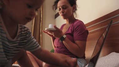 Anne, çocukla yatakta kahveyi sever. Sahne uzak çalışma dengesini ve aile hayatını yakalar. Laptop, evdeki ebeveynler için ev içi rahatlığı ve modern çalışma esnekliğini sembolize eder..