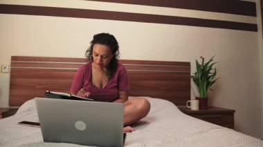 Odaklanmış bir kadın rahat bir şekilde yatağında oturur, özenle dizüstü bilgisayarı üzerinde çalışır ve bir deftere yazı yazar. Bu video, ev ortamında verimlilik ve rahatlığı vurgulayarak esnek uzaktan çalışma veya çalışmanın özünü yakalar. Temalar için ideal