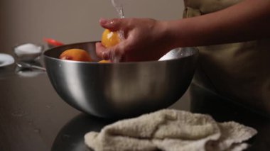 Bir insan, olgun şeftalileri metal bir kasede akan suyun altında dikkatlice yıkıyor ve onları aşçılık kullanımına karşı özenle hazırlıyor. Sahne, mutfaktaki temizlik ve hazırlığın özünü yakalıyor..