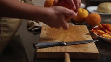 Ahşap bir tahtada şeftali soyan ve dilimleyen yakın plan bir el. Taze şeftaliler, dilimlenmiş bir tabak, ve düzgün bir şekilde yerleştirilmiş bir bıçak, pişirme ya da pişirme için bir hazırlık olduğunu gösteriyor..