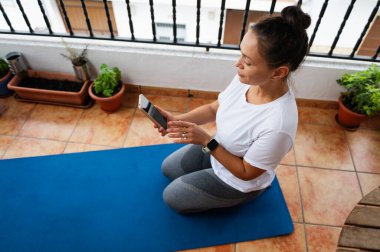 Bir kadın akıllı telefon kullanırken açık havada fayanslı bir balkonda mavi bir yoga minderinde oturuyor. Bitki saksıları alanı süsler, egzersiz veya dinlenme için sakin bir ortam önerir.