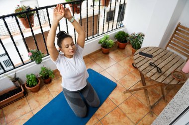 Bir kadın, yeşil saksılarla çevrili, rahat bir balkon alanında mavi paspasının üzerinde yoga yapıyor. Serene kurulum bir mobil aygıt ile bir tablo içerir, ideal bir sağIık alanı oluşturur.