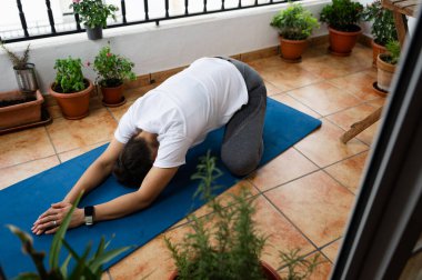Yoga pozu veren bir kadın, bitkilerle çevrili fayanslı bir balkonda, sükunetin tadını çıkarıyor..