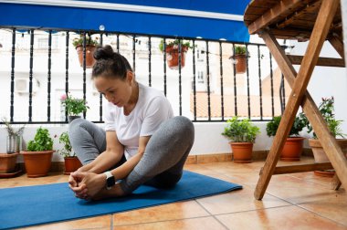Bir kadın, dingin bir balkon ortamında yoga yapıyor. Etrafı saksı bitkileri ve doğal ışıklarla çevrili. Esneklik ve rahatlamaya odaklanıyor..
