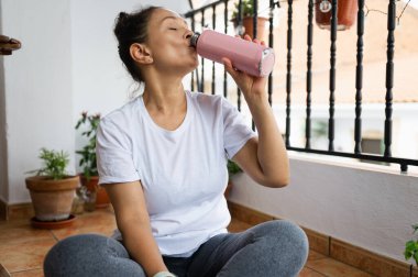 Rahat bir birey fayanslı bir balkonda bağdaş kurup ferahlatıcı pembe bir içkinin tadını çıkarıyor. Güneşli çevre, saksı bitkileri ve günlük kıyafetler huzurlu ve sağlıklı bir ortam yaratır..