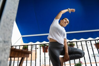 Bir kadın balkonda yoga pozisyonunda uzanıyor, açık havada açık mavi bir gölgeliğin altında spor yapıyor. Spor kıyafetleri giyiyor, sağlık, rahatlama ve farkındalık temasını vurguluyor..