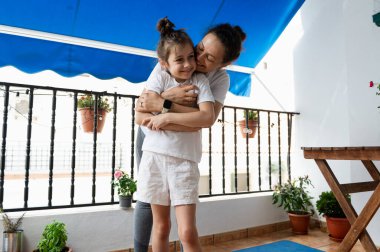 Çiçeklerle süslenmiş sıcacık bir avluda kızını kucaklayan bir anne arasında hassas bir an. Güneş ışığı, bu huzurlu ve iç açıcı ortamda paylaştıkları neşeli bağı güçlendirir..