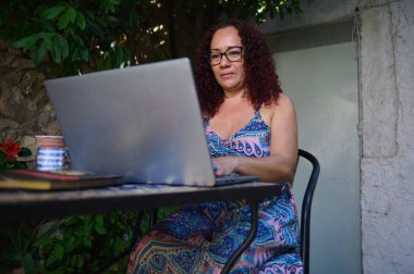 Olgun bir kadın dışarıda dizüstü bilgisayarıyla çalışır. Doğal veranda ortamında huzurlu ve üretken bir ortamın tadını çıkarır. Uzaktan çalışma, çevrimiçi iş ve rahat çalışma alanları kavramları için harika..