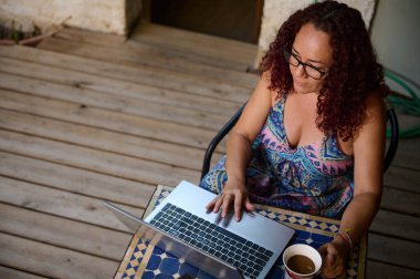 Bir kadın verandada uzaktan çalışmaktan hoşlanır. Doğa ile çevrili ve dizüstü bilgisayarını kullanarak, üretkenliği sakin bir açık hava ortamı ile birleştiriyor, odak ve rahatlamayı geliştiriyor..