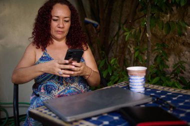 Odaklanmış bir kadın dışarıda oturuyor, akıllı telefonunu kullanıyor, yanında dizüstü bilgisayarı ve kahvesi var. Çevresi yeşilliklerle çevriliyken, huzurlu bir andan zevk alıyor, davet edici bir ortamda iş ve rahatlığı dengeliyor..