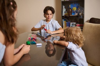 Küçük çocuklar bir masanın etrafında oturuyorlar, sanat malzemeleri ve aletlerle uğraşıyorlar, yaratıcılığı teşvik ediyorlar. Raflar ve kitaplarla dolu rahat bir oda arka planında, yetiştirme ve aile odaklı bir atmosfer vurgulanıyor.