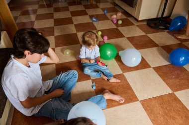 Genç bir anne ve çocuğu evin içinde neşeli bir anın tadını çıkarıyorlar, kareli zeminde renkli balonlarla çevrili, günlük bir aile ortamında sıcaklığı ve bağ kurmayı teşvik ediyorlar..