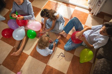 Bir aile fayanslı bir zeminde toplanıyor, balon enflasyonu ve el işi faaliyetleri, yaratıcılığı ve bağ kurmayı teşvik ediyor. Parlak renkler ve doğal ışık, neşeli ve iç açıcı bir anı vurgular..