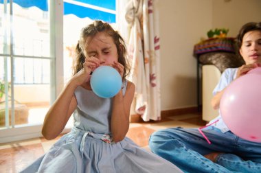 İki çocuk birlikte balon şişirerek eğlenceli bir etkinliğe katılıyorlar. Doğal ışıklandırma, oyun zamanı boyunca keyif ve yaratıcı enerjilerini ön plana çıkararak konforlu alanı aydınlatır..