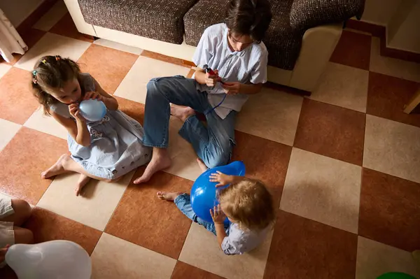 Üç çocuk kapalı bir zeminde rahat mobilyalarla oyun oynayıp balon şişiriyor. İçerde neşeli ve eğlenceli bir atmosfer yakalamak, çocukluk mutluluğu ve eğlencesini sergilemek..