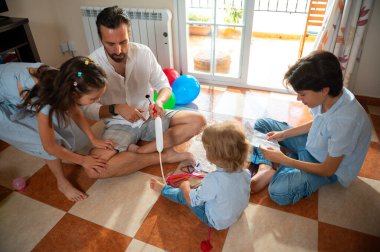 Sıcak bir oturma odasında toplanan bir aile, yaratıcı aktiviteleri coşku ve sevgiyle gerçekleştiriyor. Balonlar ve oyuncaklar boşluğu dolduruyor, eğlenceli ve iç açıcı bir aile atmosferini vurguluyorlar..