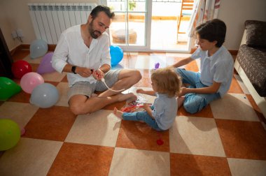 Şefkatli bir baba iki çocukla fayanslı zeminde oturup balon oyunu oynar. Sahne neşe ve aile bağlarını yansıtıyor. Parlak, sıcak güneş ışığı altında bir oturma odasında geçiyor..