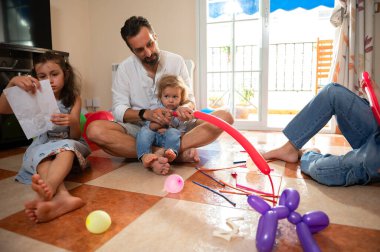 Bir babanın çocuklarıyla evde eğlenceli aktiviteler yaptığı bir aile bağı anı. Kapalı alanda balonlar ve el sanatları malzemeleri bulunur. Neşeli ve sıcak bir aile ortamı yayarlar..