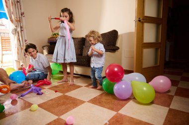 Üç çocuk güneşli, rahat bir odada renkli balonlarla oynuyorlar. Neşeli atmosfer kutlama, takım çalışması ve eğlence öneriyor. Yakın aile ortamında neşe ve yaratıcılığı somutlaştırıyor..