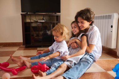 Üç çocuk oturma odasının fayanslı zemininde oturmuş oyuncaklarla oynuyor ve gülüyorlar. Etkileşimlerinin sıcaklığı ve enerjisi davetkar, eğlenceli ve aile odaklı bir atmosfer yaratır..