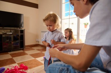 Bir aile güzel ışıklandırılmış, rahat bir oturma odasında balondan hayvan yapmak için kaliteli zaman geçirir. Çocuklar renkli balonları şekillendirmeye, yaratıcı ve eğlenceli aktivitelere birlikte odaklanırlar..