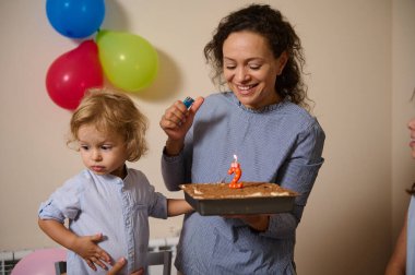 Bir anne çocuğunun ikinci doğum günü kutlaması için elinde mum olan bir pasta tutar. Etrafı balonlarla çevrilidir..