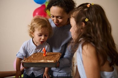 Bir anne ve iki çocuğu birlikte doğum gününü kutluyorlar. Oğlan, kız izlerken pastanın üzerine tek bir mum üflüyor. Şenlikli dekorasyonlar bir neşe ortamı sağlar..
