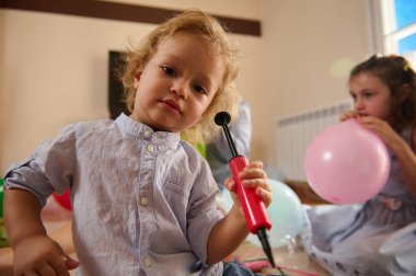 Genç bir çocuk aktif olarak balon pompası kullanırken diğer bir çocuk da rahat bir ortamda balon şişiriyor. Görüntü sıcaklığı, eğlenceyi ve aile yakınlığını yayar..