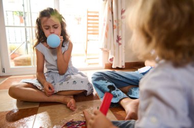 Bir grup çocuk içeride eğleniyor, balon şişiriyor ve yerde oturuyor. Güneş ışığı içeri akar, oturma odasında neşeli ve rahat bir atmosfer yaratır..