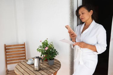 Kadınlar verandada sakin bir sabah geçirir, akıllı telefonunu kullanır ve dinlenir. Ahşap bir masanın üzerine kurulmuş bir kahve ve bitkiler sıcaklık katıyor. Sahne rahatlık, rahatlık ve refahı iletir..