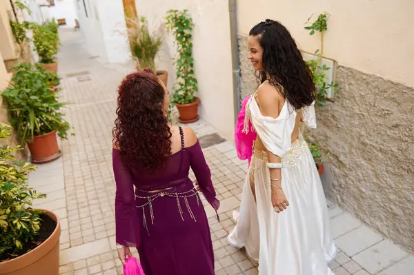 Canlı mor ve beyaz giyinmiş iki kadın huzurlu, çiçekli bir sokakta yürüyorlar. Zarif elbiseler giyip mutluluk saçıyorlar. Büyüleyici bir mimariyle çevrili güzel bir günün tadını çıkarıyorlar..