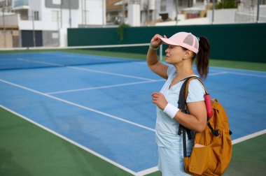 Mavi tenis kortunun yanında kep ve sırt çantası takan bir tenisçi. Parlak güneş ışığı altında sakin ve düşünceli görünüyor. Hafif bir spor kıyafeti giyiyor..