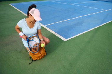 Spor elbiseli bir kadın açık hava sahasında tenis oynamaya hazırlanıyor. Ekipmanlarını sırt çantasında düzenliyor. Canlı yeşil ve mavi mahkeme eğlence ya da profesyonel spor sahnesini hazırlıyor.