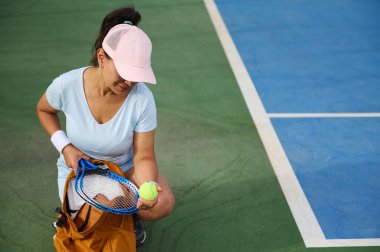 Spor kıyafetleri giymiş bir bayan tenisçi açık hava tenis kortunda antrenman yapmaya hazırlanıyor. Maçtan önce raketini ve topunu kontrol ediyor. Odaklanma ve kararlılık gösteriyor..