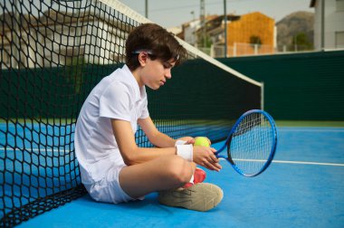 Beyaz elbiseli genç bir tenis atleti canlı bir mavi tenis kortunda elinde raket ve tenis topları tutarken ağın kenarında dinleniyor. Kompozisyon spor ve kararlılığı vurguluyor.