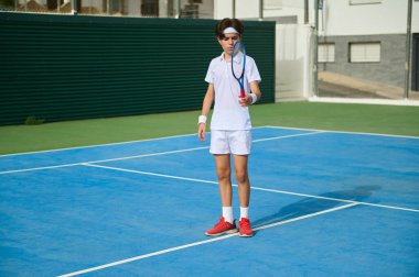 Genç bir atlet açık hava sahasında tenis çalışıyor. Etkinlikte beceri ve konsantrasyon üzerinde durulurken, destekleyici aksesuarlar performansı artırıyor. Kararlılık ve sporun dinamik bir sahnesi.