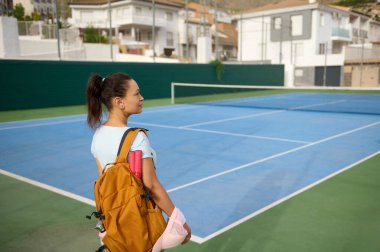 Genç bir kız tenis kortunun yanında duruyor, sırt çantası ve tenis teçhizatı taşıyor. Dekor, bir açık hava mahkemesi ve kentsel çevreyi de içeriyor. Spor ve yaşam tarzı temaları için ideal.