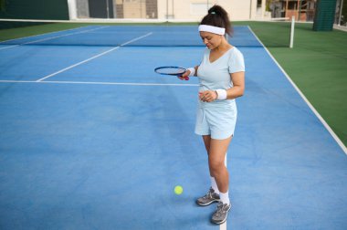 Açık mavi atletizm kıyafeti giyen bir kadın tenis yeteneklerini açık hava mavi kortunda iyi bir ışık altında çalışır..